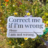 A die-cut white sign with black text reading 'Correct me if I'm wrong Phrase I am not wrong.' placed in a garden among flowers and greenery, with a metal stake holding it.