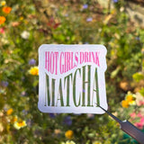 A die-cut sticker with the text 'HOT GIRLS DRINK MATCHA' in pink and green lettering on a white background, held by a hand using tweezers against a blurred garden backdrop of green foliage and small yellow flowers.
