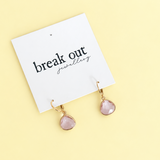 A pair of gold-tone drop earrings with faceted light pink teardrop gemstones, displayed on a white card that reads 'break out jewellery' against a pale yellow background.