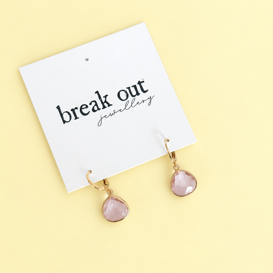 A pair of gold-tone drop earrings with faceted light pink teardrop gemstones, displayed on a white card that reads 'break out jewellery' against a pale yellow background.