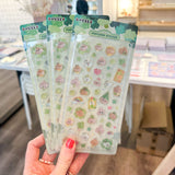 A hand with red nail polish holds three packages of cute, pastel-colored popcorn-themed stickers in a shop setting with wooden floors and shelves in the background.
