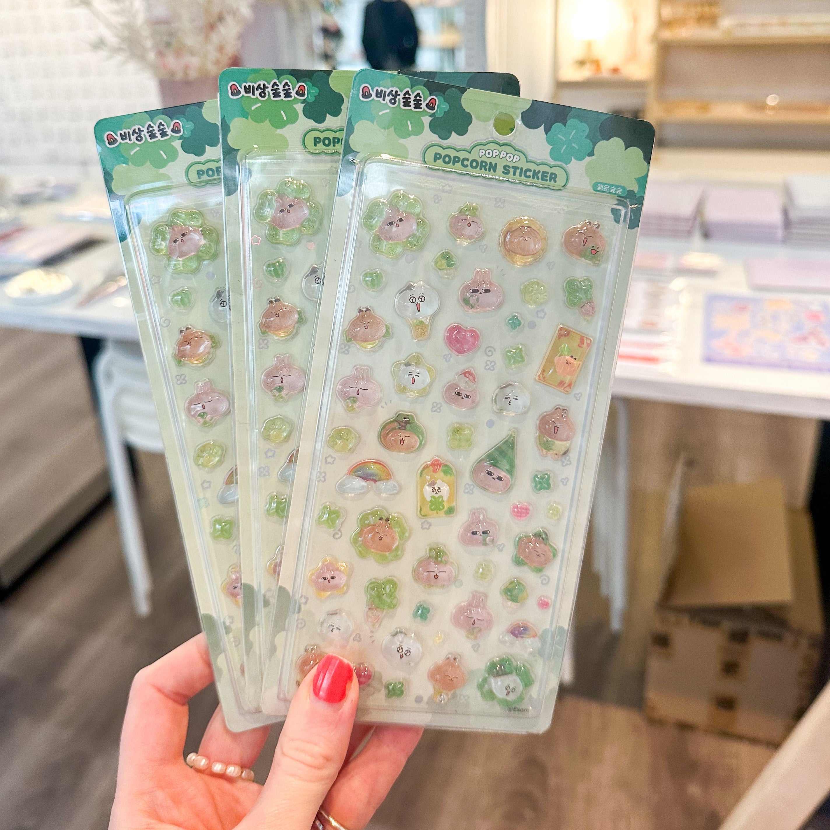 A hand with red nail polish holds three packages of cute, pastel-colored popcorn-themed stickers in a shop setting with wooden floors and shelves in the background.
