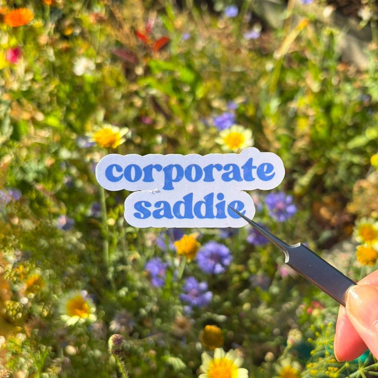A hand holds a small black spatula against a blurred garden background filled with yellow, purple, and white wildflowers. The spatula is positioned over a sticker or label that reads 'corporate saddie' in blue bubbly text on a light blue background.