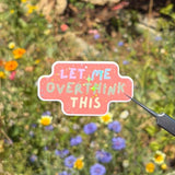 A hand holds tweezers with a small die-cut sticker that reads 'LET ME OVERTHINK THIS' in colorful lettering on a pink background, set against a blurred outdoor garden with wildflowers.