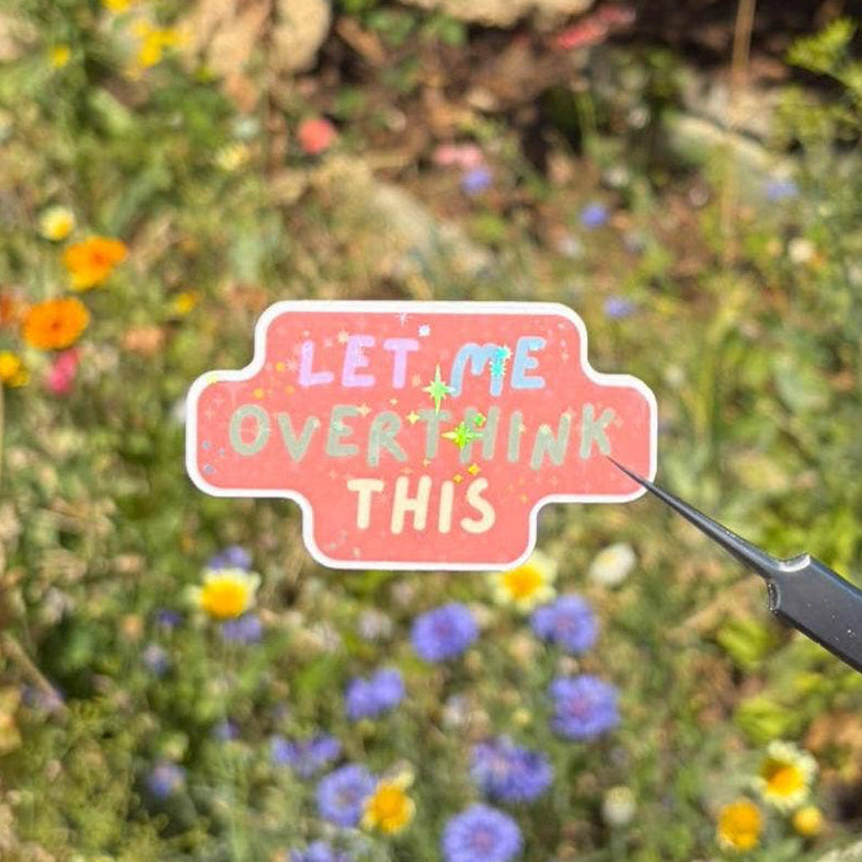 A hand holds tweezers with a small die-cut sticker that reads 'LET ME OVERTHINK THIS' in colorful lettering on a pink background, set against a blurred outdoor garden with wildflowers.