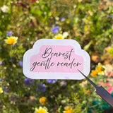 A hand holds a small die-cut sticker with the phrase 'Bearest gentle reader' in black script on a pale pink background with a white border. The sticker is pointed at by a small metal tool, with a blurred outdoor flower field in the background.