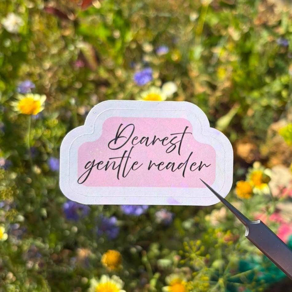 A hand holds a small die-cut sticker with the phrase 'Bearest gentle reader' in black script on a pale pink background with a white border. The sticker is pointed at by a small metal tool, with a blurred outdoor flower field in the background.