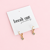 A pair of small gold hoop earrings with tiny pastel blue-green square charms, displayed on a white card that reads 'break out jewellery' against a soft pink background.