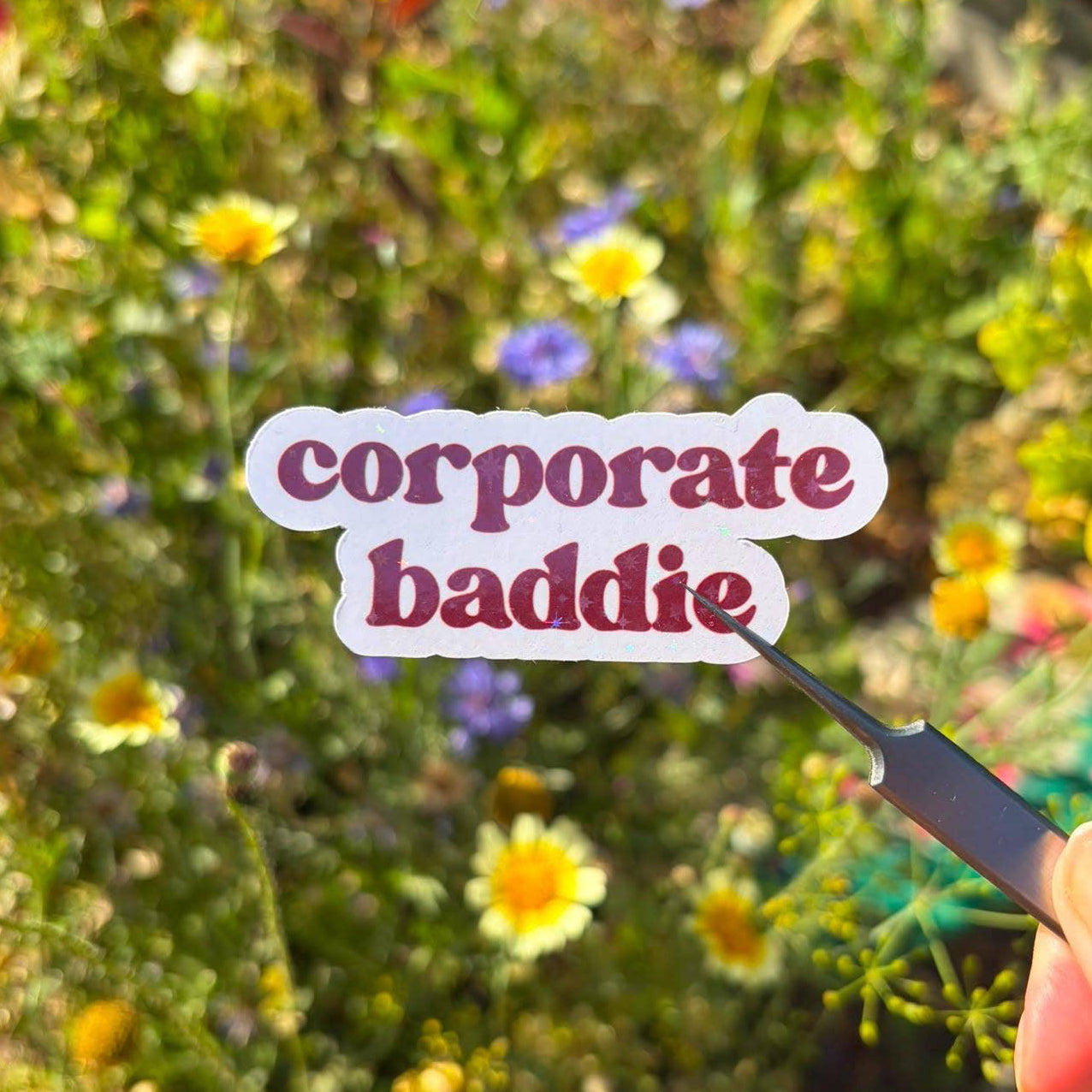 A die-cut sticker with the words 'corporate baddie' in dark pinkish-purple text on a white background, held by tweezers over a blurred garden of yellow and white flowers.
