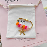 A gold-toned carabiner keyring with pink and orange beads and a starfish charm is displayed on a white fabric pouch over a pink card, with jewelry accessories arranged around it on a round table.