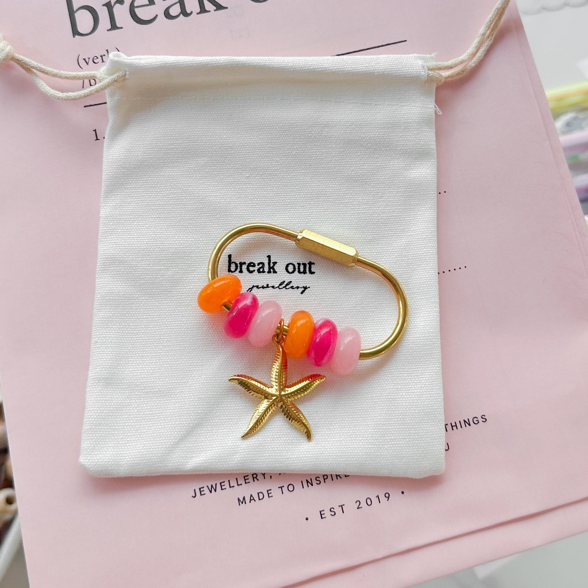 A gold-toned carabiner keyring with pink and orange beads and a starfish charm is displayed on a white fabric pouch over a pink card, with jewelry accessories arranged around it on a round table.