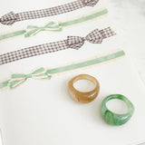 Two chunky ring-style bracelets, one translucent amber-brown and one translucent green, displayed on a white notebook page alongside ribbon samples.