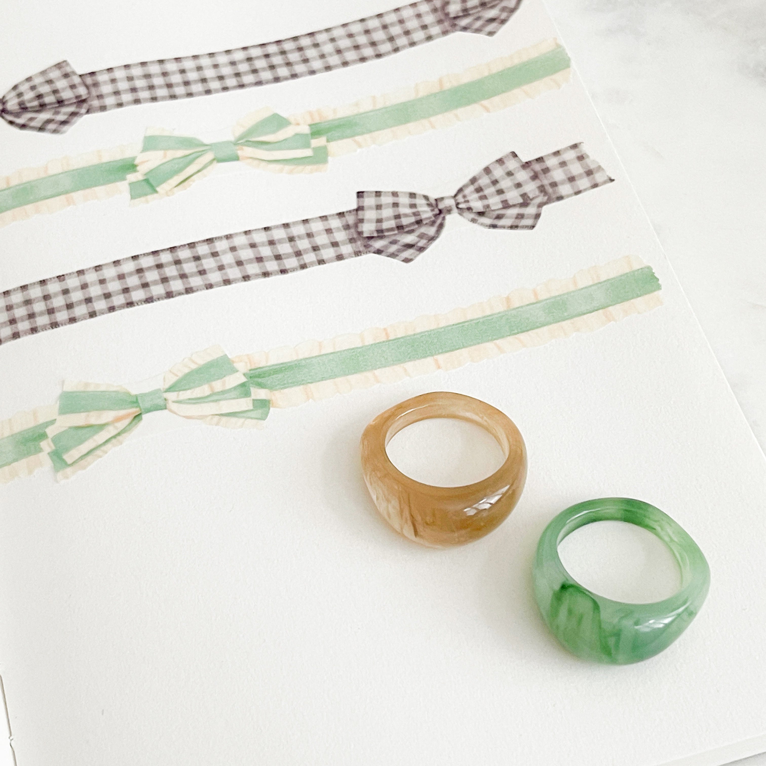 Two chunky ring-style bracelets, one translucent amber-brown and one translucent green, displayed on a white notebook page alongside ribbon samples.