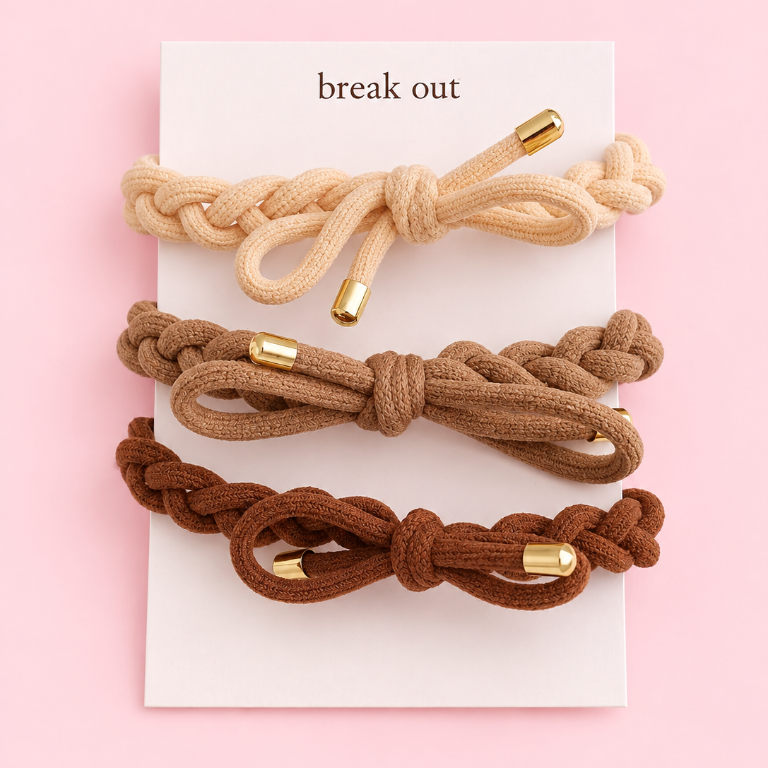 Three braided cord hair ties in beige, brown, and dark brown, each with a gold metal end cap, arranged on a white display card that reads 'break out' against a solid pink background.
